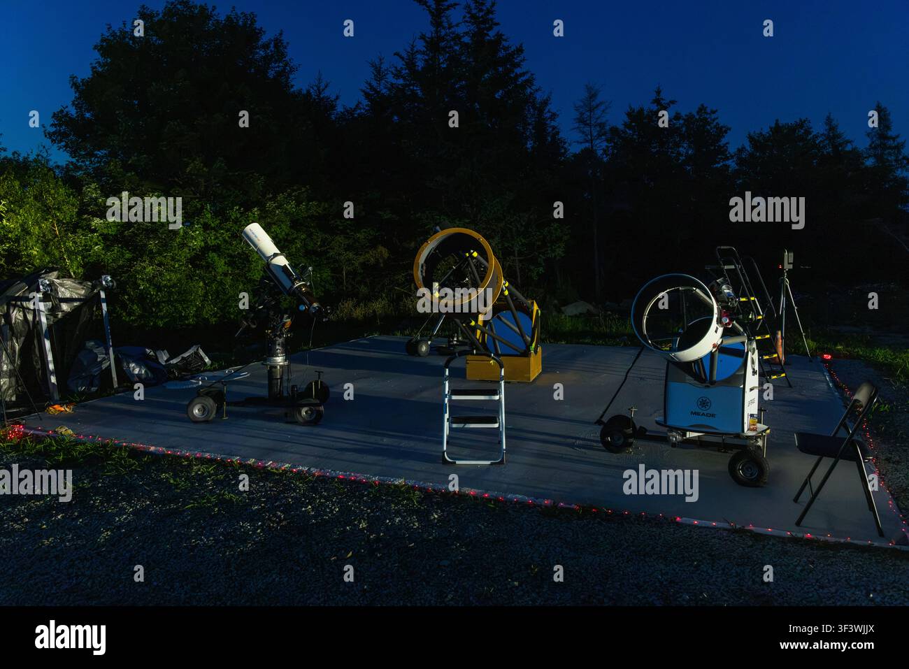 At dusk, three different telescopes stand ready for use on the open-air grounds of the Deep Sky Eye Observatory on Frotten Road in the Municipality of Argyle, Nova Scotia, Canada. Fairy lights along the edge of the platform mark the paths between the observation stations. Argyle Municipal, Nova Scotia, Canada Stock Photo