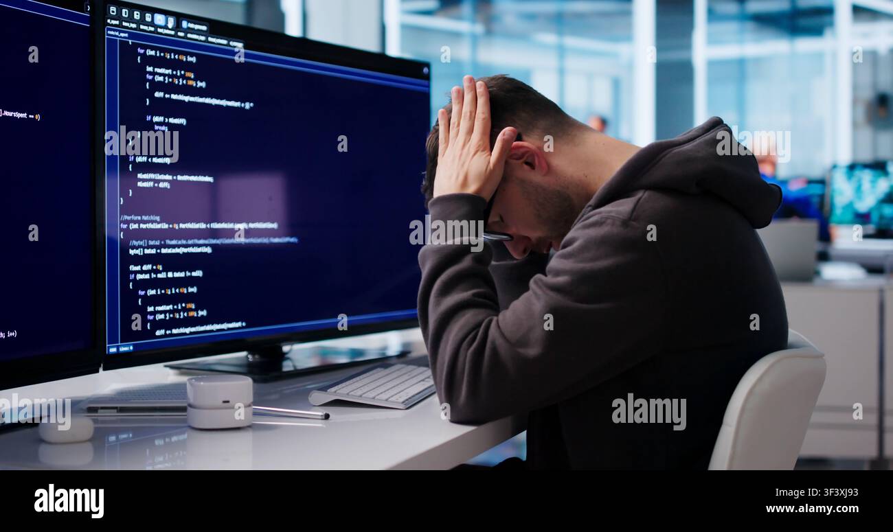 Young Software Engineer Looks Tired And Stressed While Coding At Desk. Stock Photo