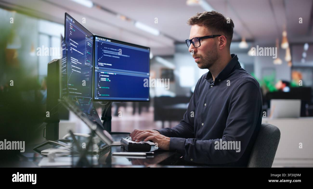 Young Software Engineer Works On Complex Algorithms Using Dual Screens At Desk. Stock Photo