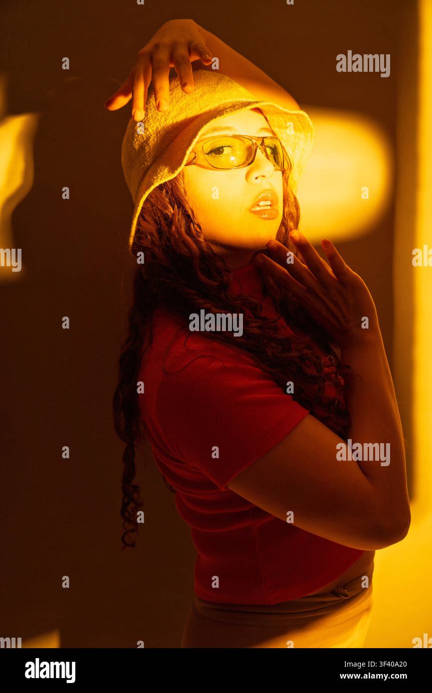 Young woman wearing a bucket hat and sunglasses posing with hands, illuminated by warm orange light Stock Photo
