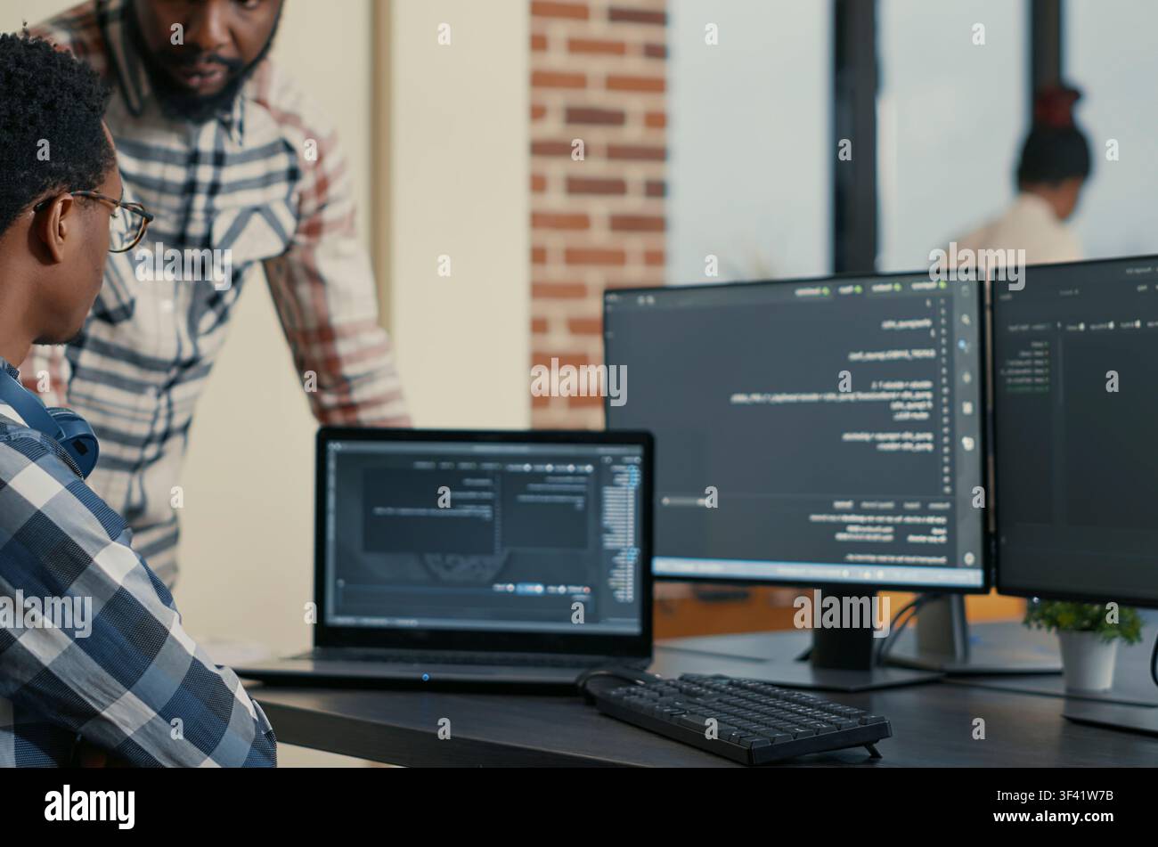 Software developer writes code on a laptop while a coworker asks for advice, showing programming teamwork and cloud computing collaboration. Stock Photo