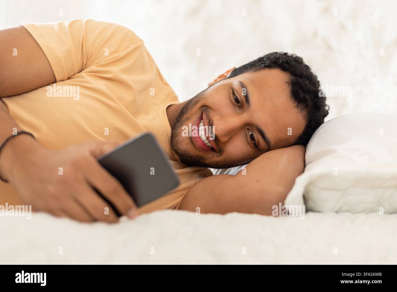 Smiling Young Black Man Using Smartphone While Lying On Bed, Relaxed Indoor Setting Stock Photo