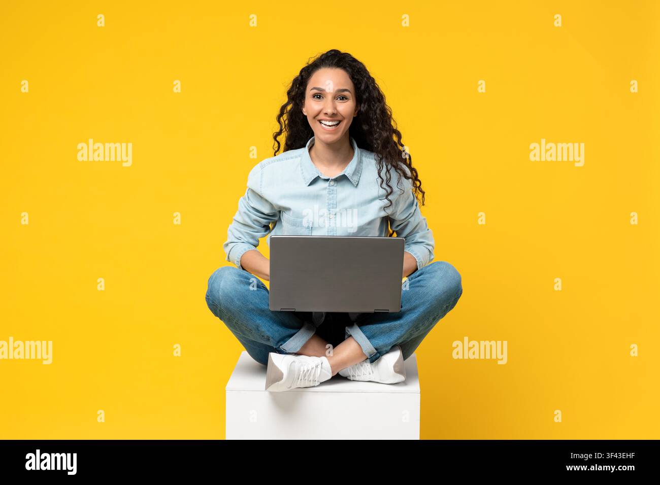 Smiling Young Mixed Race Woman Working On Laptop, Sitting On Cube, Yellow Background Stock Photo