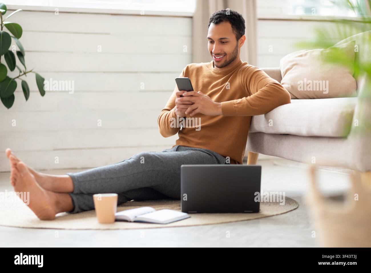 Smiling Young Hispanic Man Using Smartphone While Sitting On Floor In Cozy Living Room Stock Photo