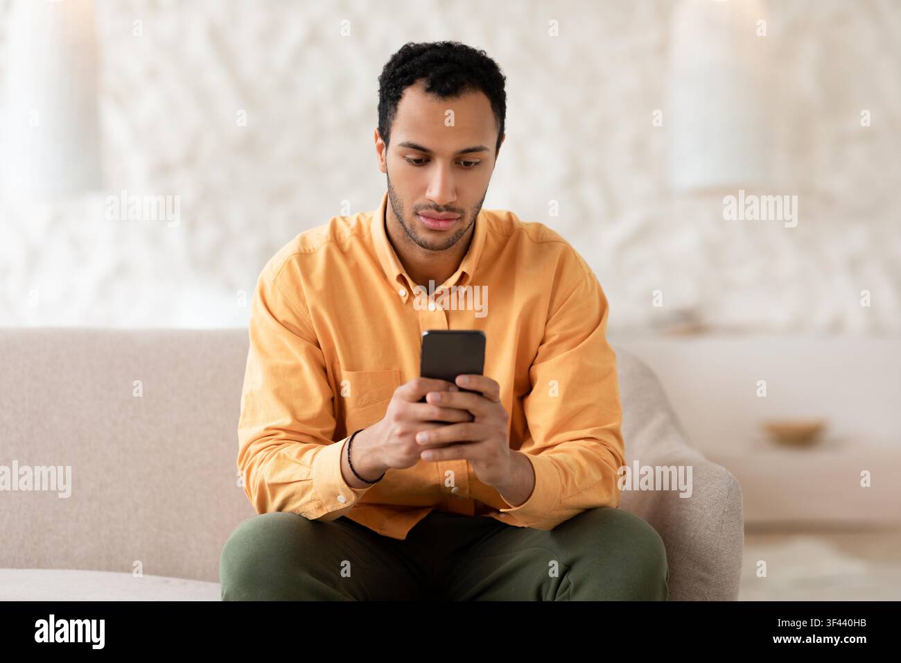 Focused Young Black Man Using Smartphone While Sitting On Couch, Indoor Setting Stock Photo