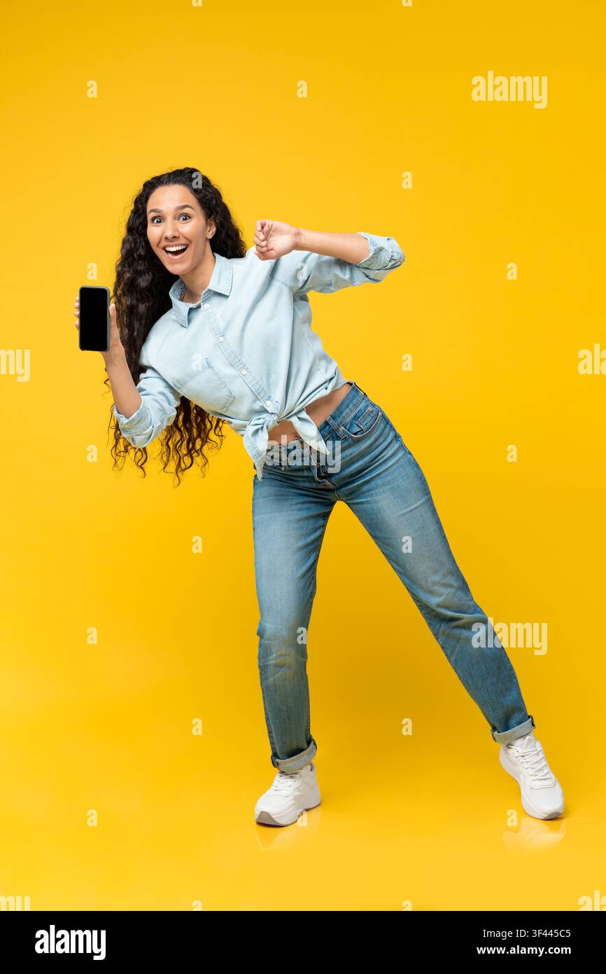 Happy Young Hispanic Woman Holding Smartphone And Dancing, Yellow Background, Mockup Stock Photo