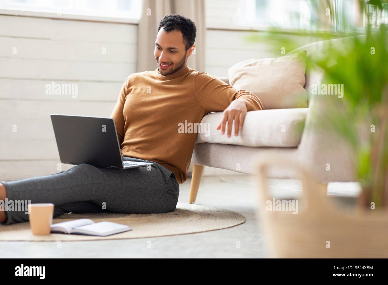 Smiling Young Black Man Working On Laptop While Sitting On Floor At Home, Professional Concept Stock Photo