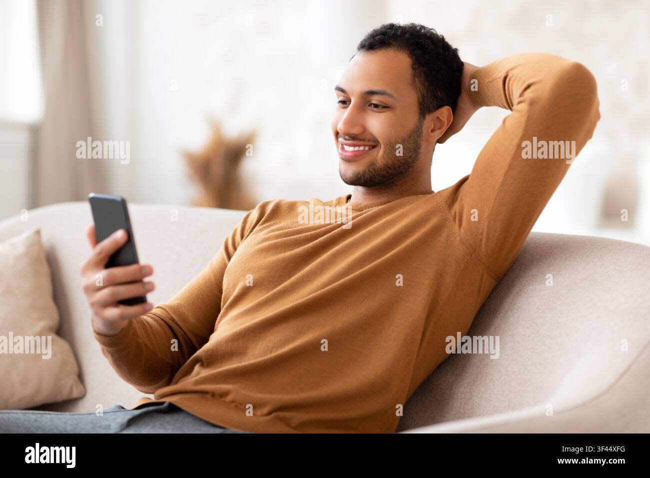 Smiling Young Man Relaxing On Sofa While Looking At Smartphone At Home Stock Photo