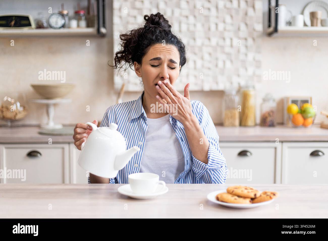 Tired Young Woman Yawning While Pouring Coffee In Kitchen, Morning Routine Stock Photo