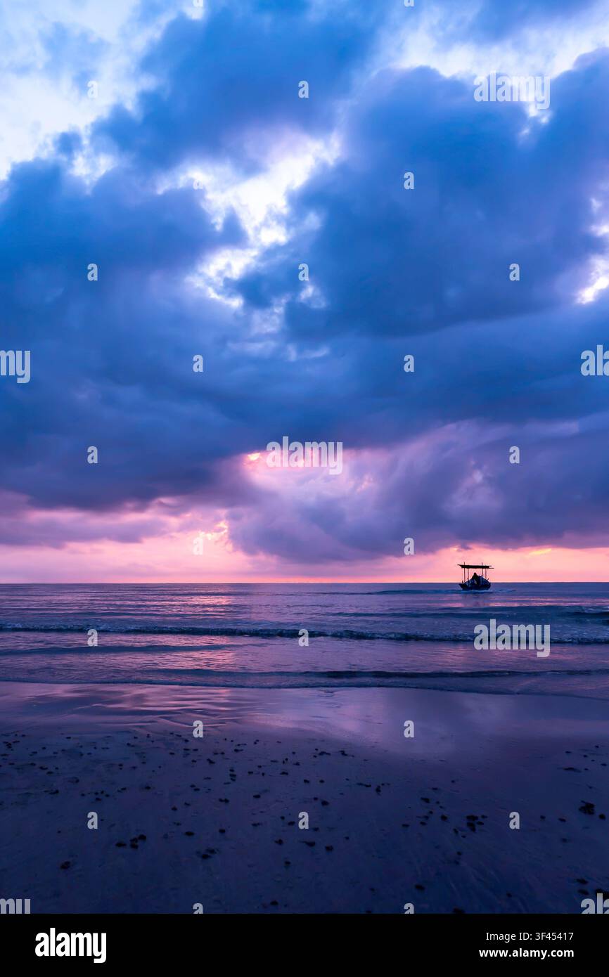A lone boat sails across a tranquil ocean under a dramatic sky painted with vibrant purple, blue, and pink hues during a breathtaking sunrise. Reflect Stock Photo