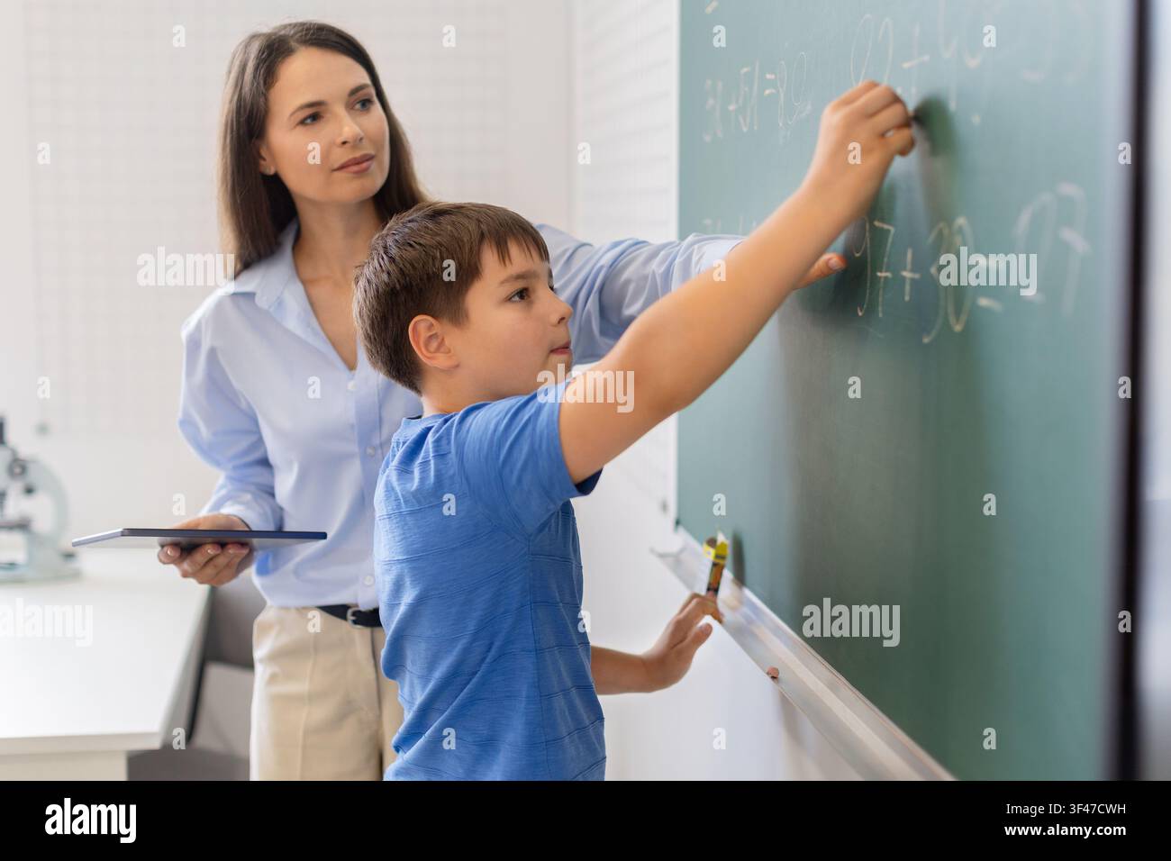 Focused schoolboy writing math equation on chalkboard while teacher holding tablet Stock Photo