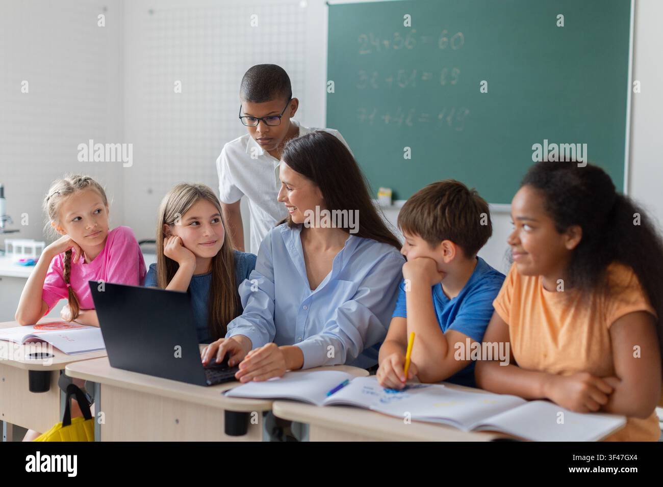 Female teacher using laptop while discussing classwork with diverse group of schoolchildren Stock Photo