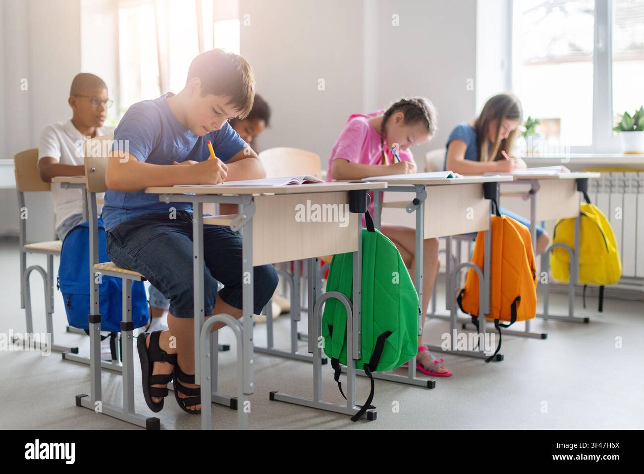 Diverse schoolchildren completing written assignments at individual desks in bright classroom Stock Photo
