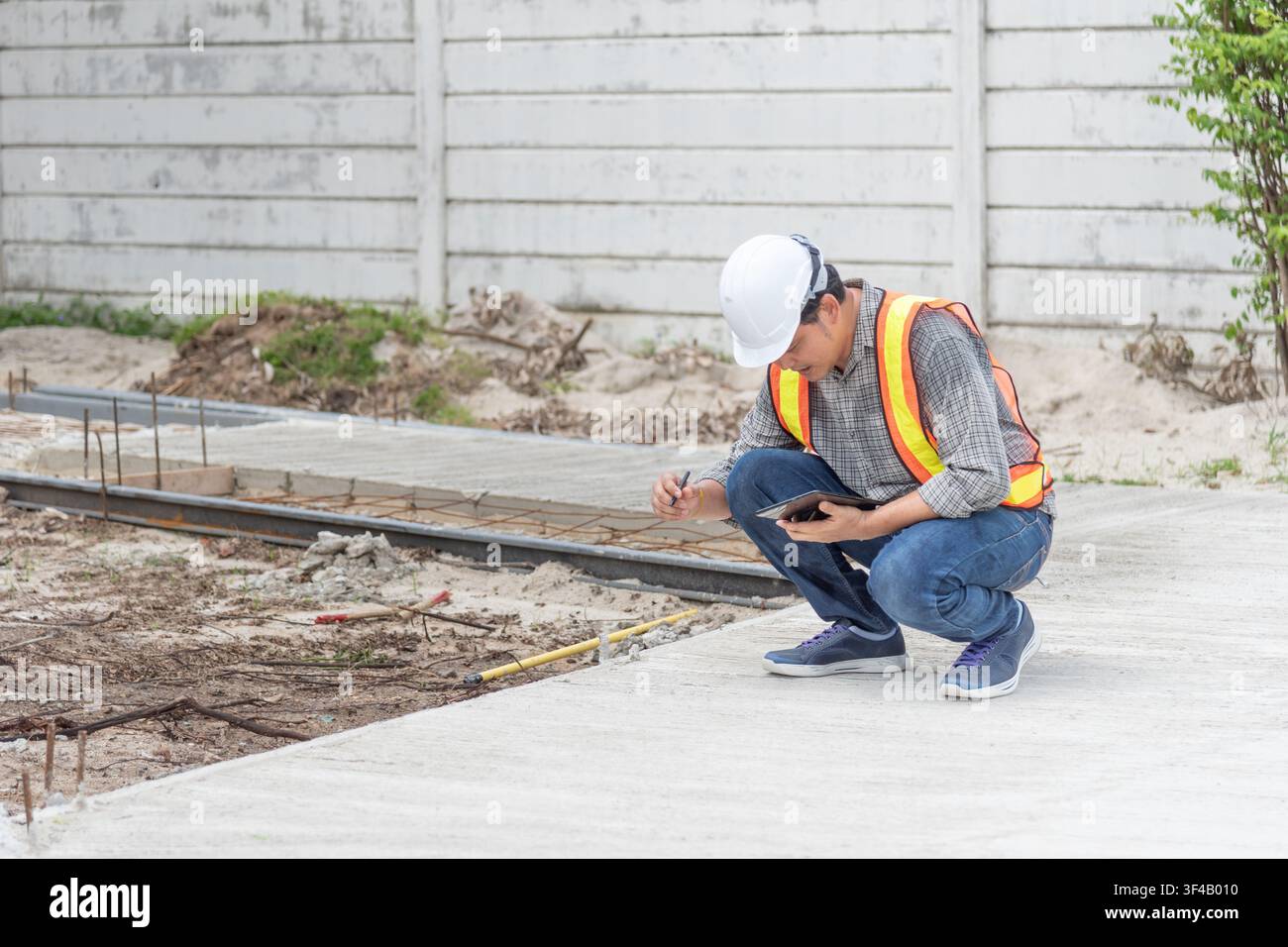 Construction engineer inspecting concrete pavement at a building site using a tablet and taking notes while checking quality safety and project progre Stock Photo