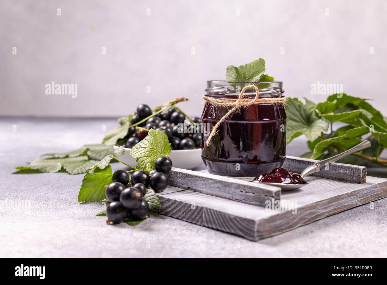 Black currant jam in a glass jar, adorned with twine, sits on a rustic wooden tray beside a bowl of fresh black currants and green leaves, with a spoo Stock Photo