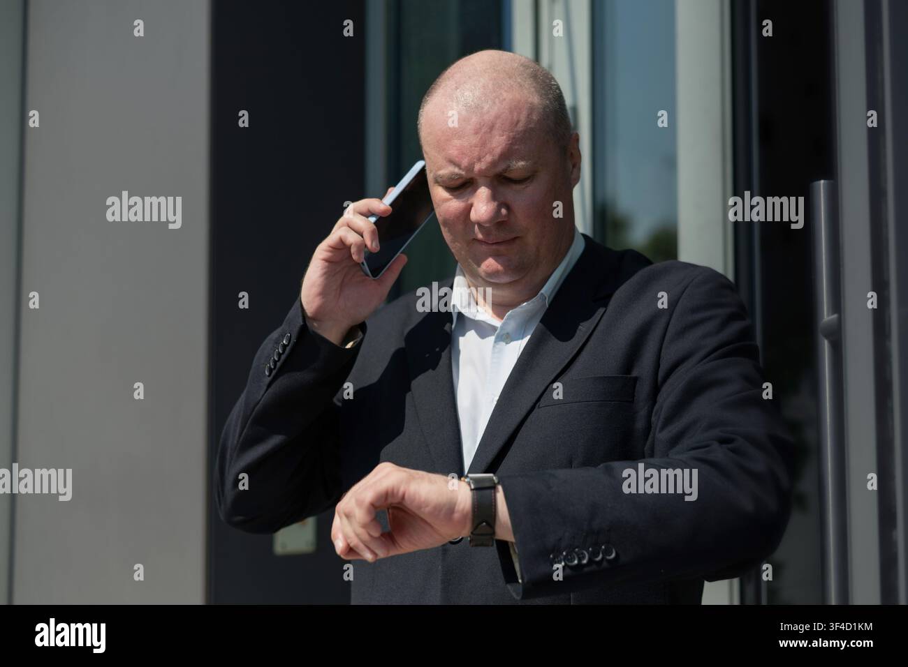 Businessman checking smartwatch while talking on smartphone outside office Stock Photo
