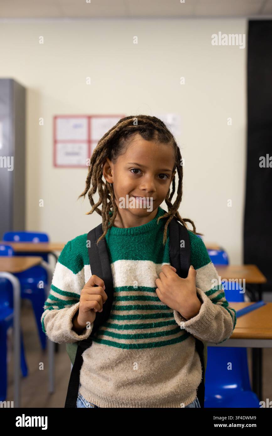 African American schoolboy standing at classroom front holding black backpack in striped sweater Stock Photo