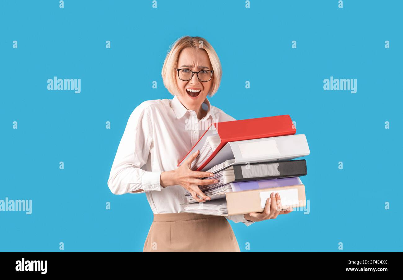Frustrated office worker overloaded with papers, holding folders of documents on blue studio background Stock Photo