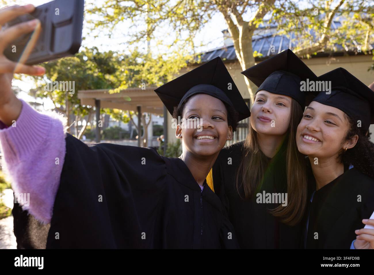 Diverse teen female students taking selfie on campus with smartphone, caps, gowns, tassels Stock Photo