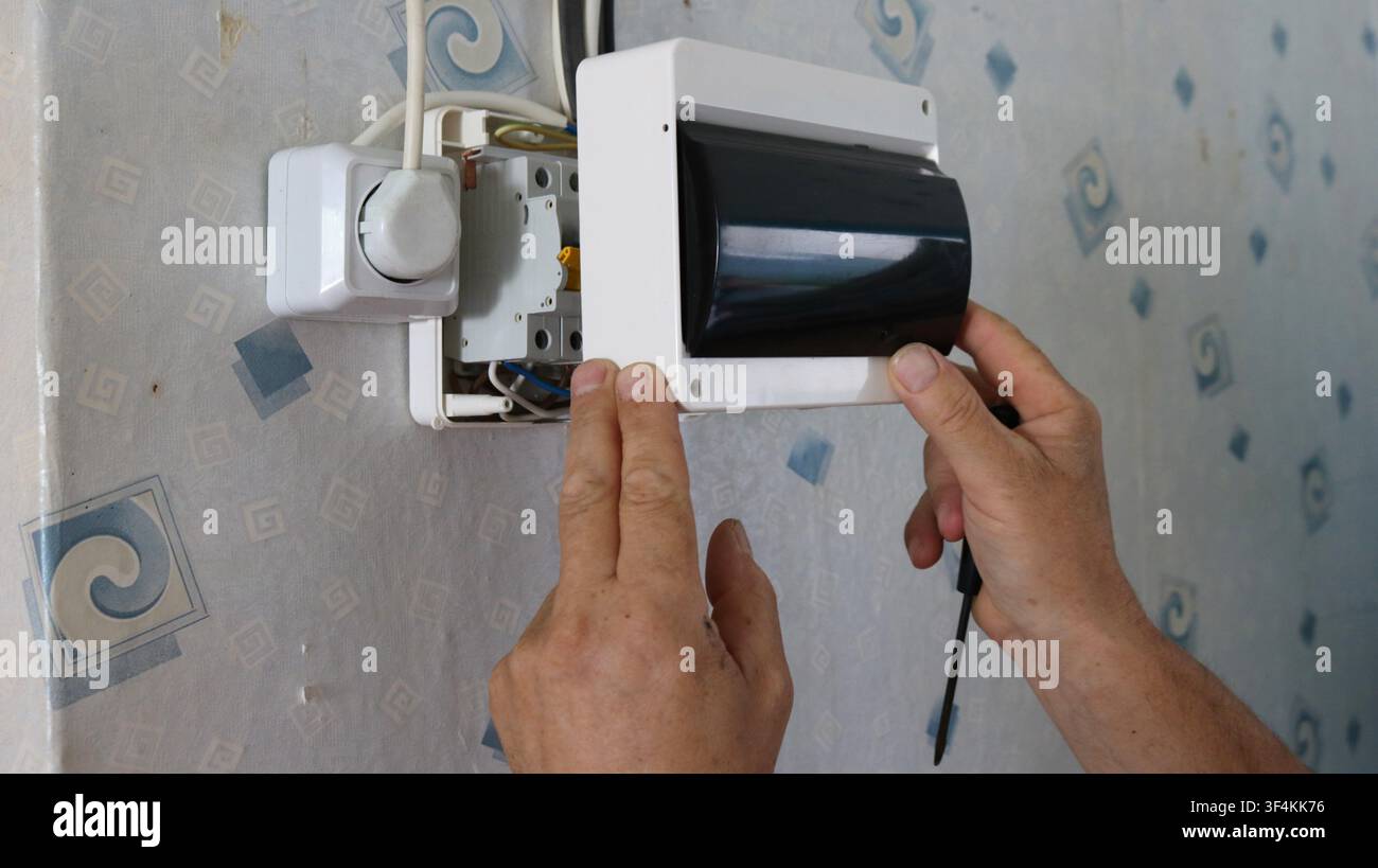 installing an electrical panel on a wallpapered wall. Hands position the white plastic housing with a dark protective cover over the circuit breakers Stock Photo