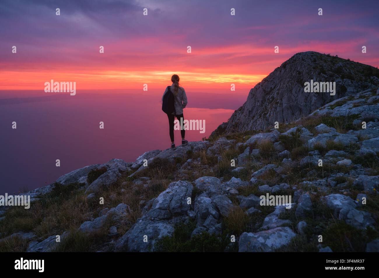 Woman with backpack standing on rocky mountain top at sunset Stock Photo