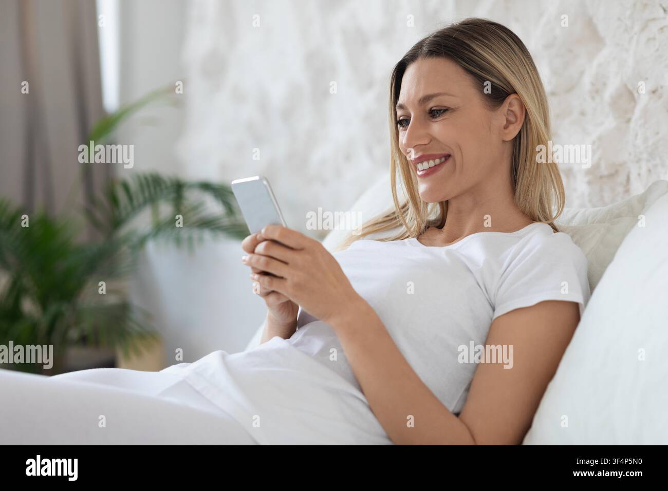 Smiling Young Woman Using Smartphone While Lying On Couch At Home, Lifestyle Concept Stock Photo