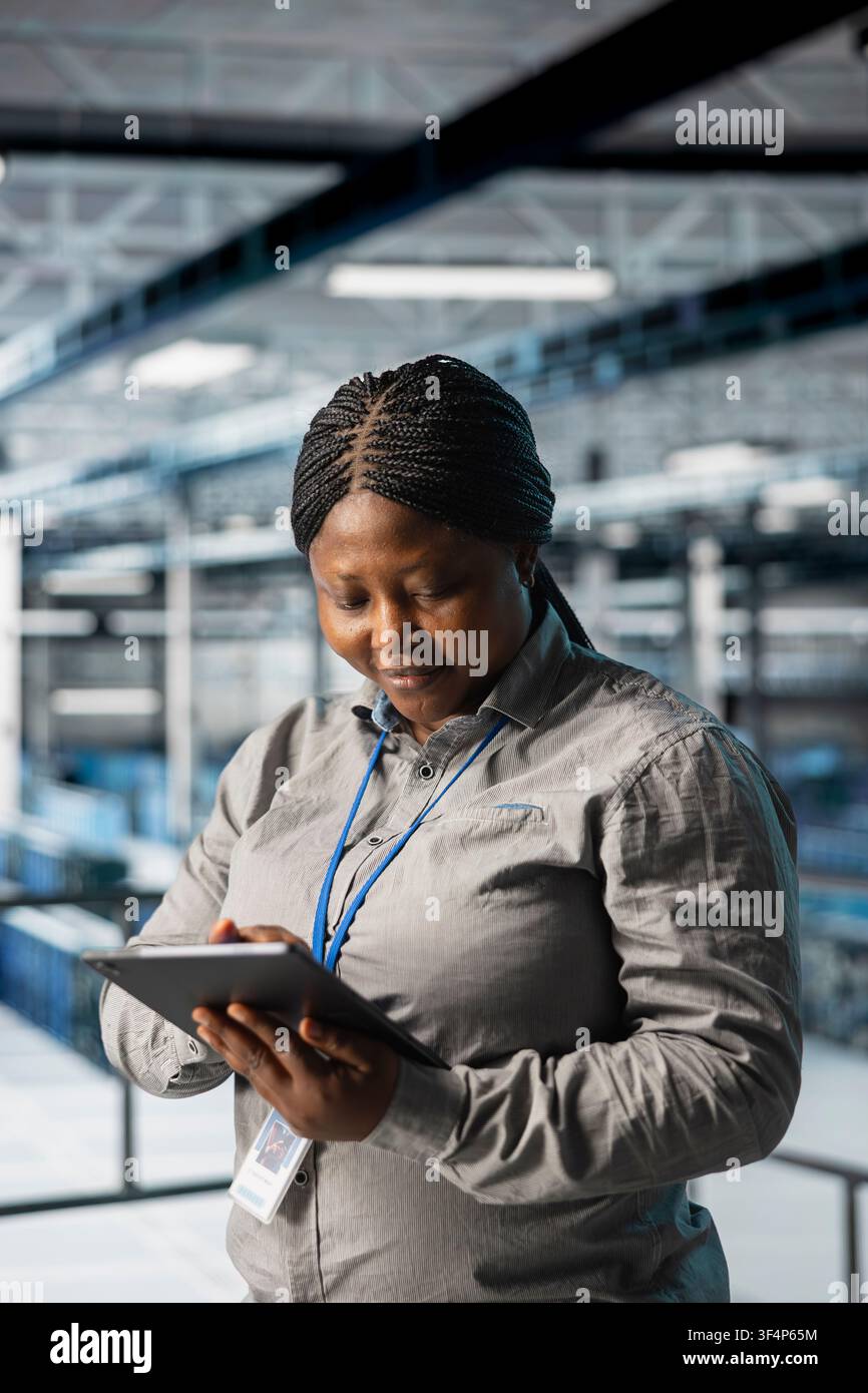 Female programmer in data center using tablet to manage software updates for system stability. African american woman in server room optimizing performance with device, configuring infrastructure Stock Photo