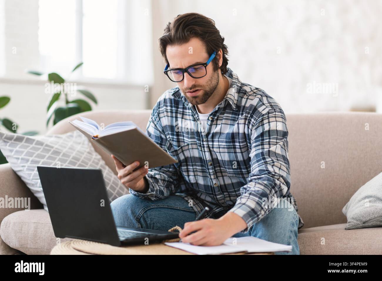 Focused Young Man Reading Book While Working On Laptop At Home, Study Concept Stock Photo
