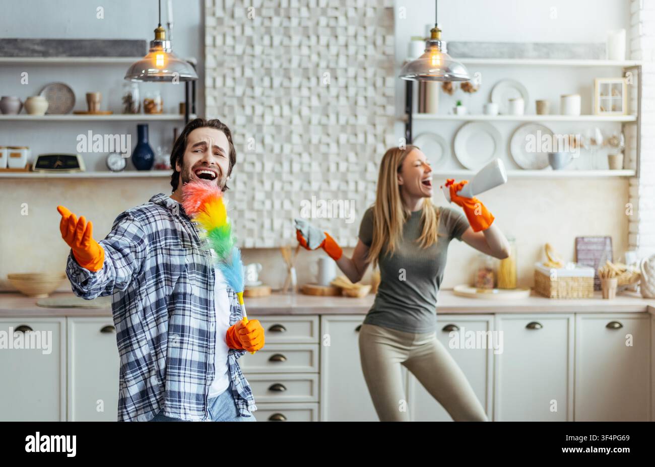 Happy Young Couple Cleaning Kitchen Together, Man Singing With Feather Duster, Woman Dancing Stock Photo