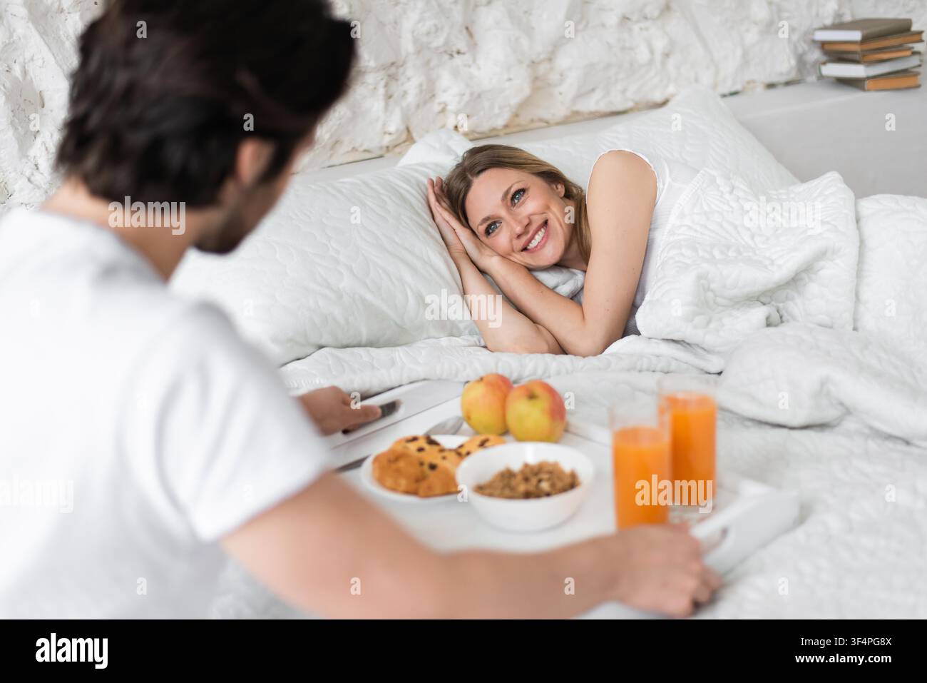 Happy Young Woman Smiling At Partner With Breakfast In Bed, Cozy Bedroom Setting Stock Photo