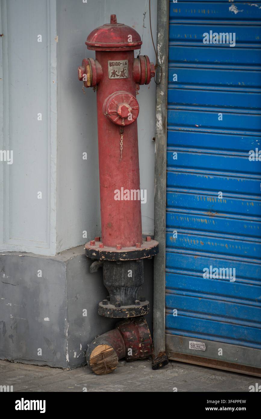 A weathered red fire hydrant stands vertically against a light gray concrete wall, to the left of a corrugated blue metal rolling shutter door. The hy Stock Photo