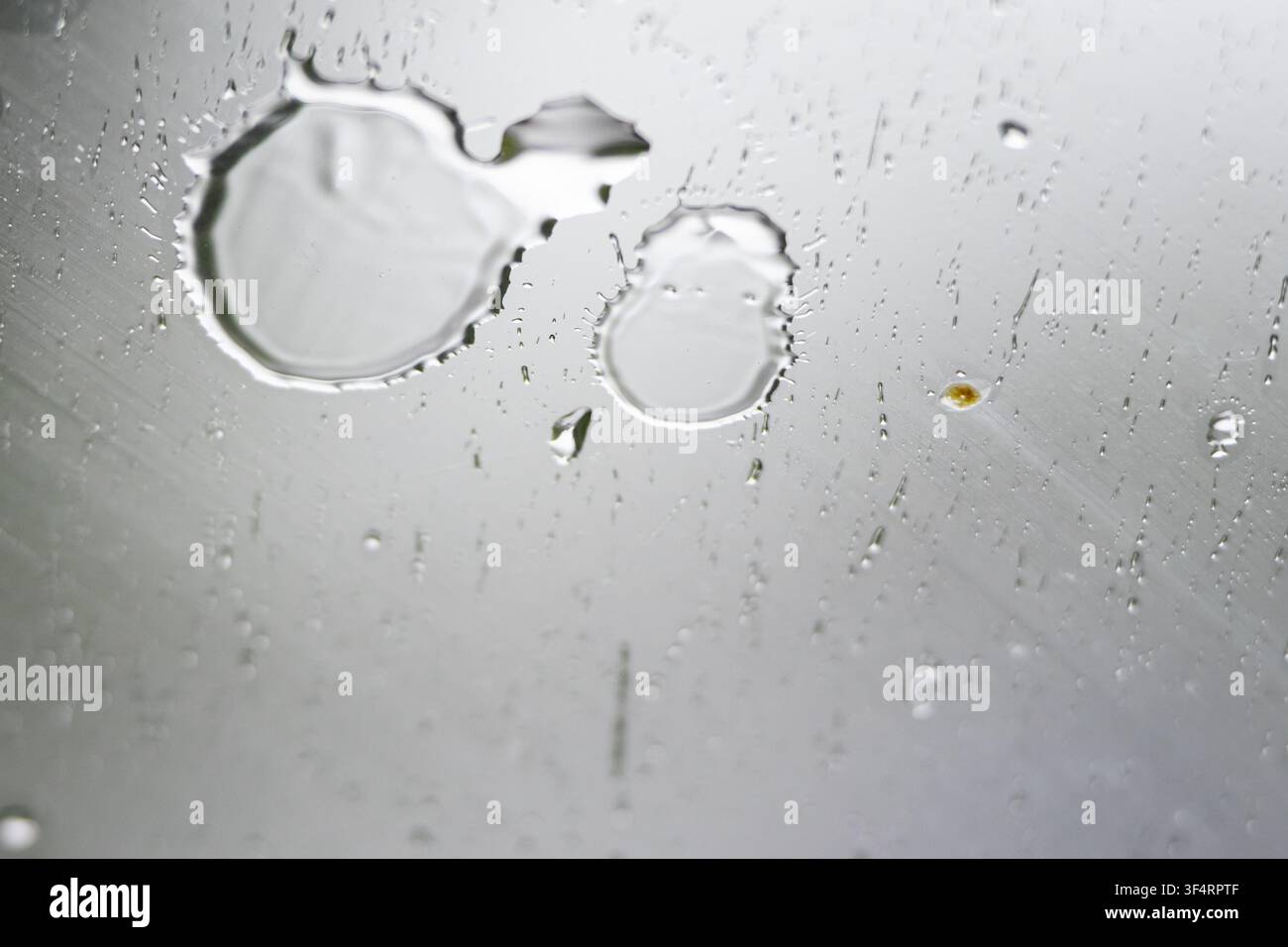 Macro view of rain droplets on smooth glass. Fine streaks slide downward as gravity works. Soft gray light fills the scene creating a calm abstract mo Stock Photo