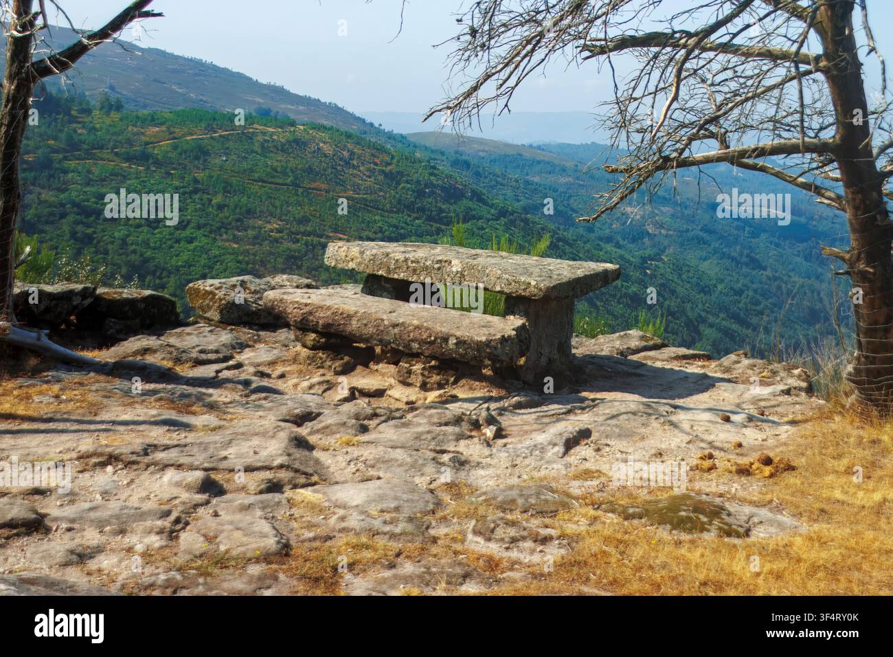 Empty stone picnic bench on a high rocky viewpoint overlooking scenic green valleys and hills during a bright summer day, outdoor travel concept. Stock Photo