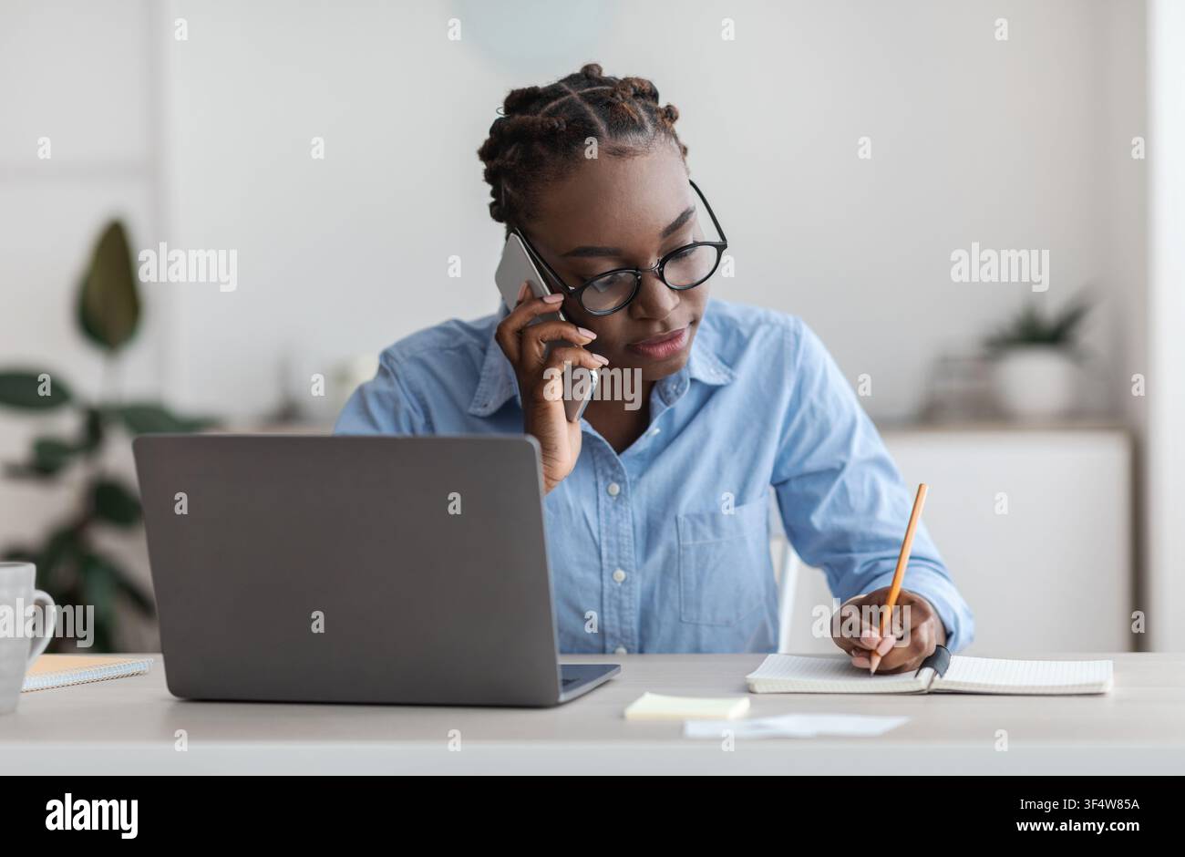 Black Female Office Worker Talking On Cellphone With Client And Taking Notes Stock Photo