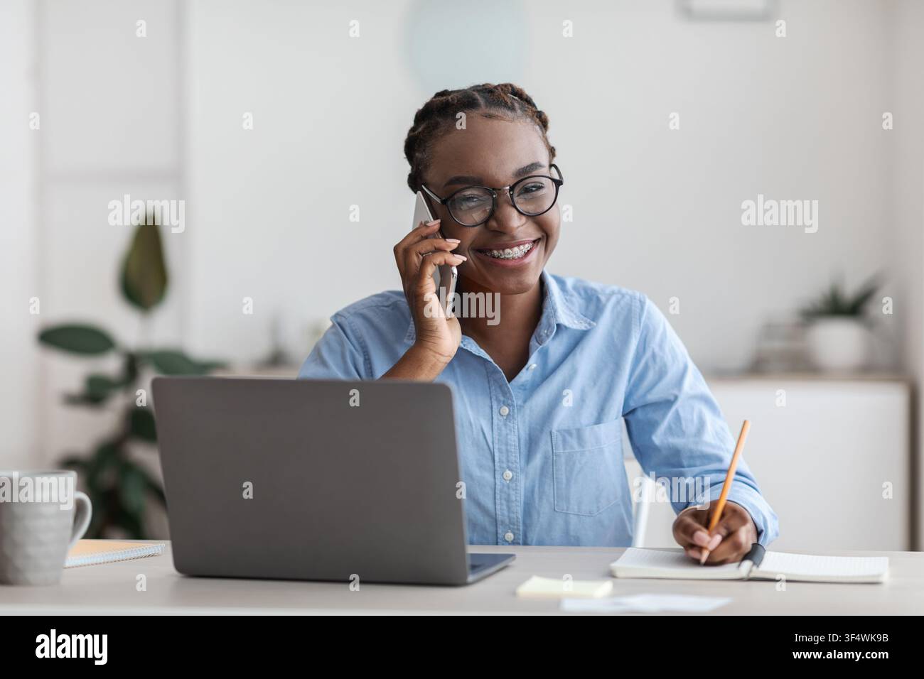 Office Trainee. Black woman talking on cellphone and taking notes at workplace Stock Photo