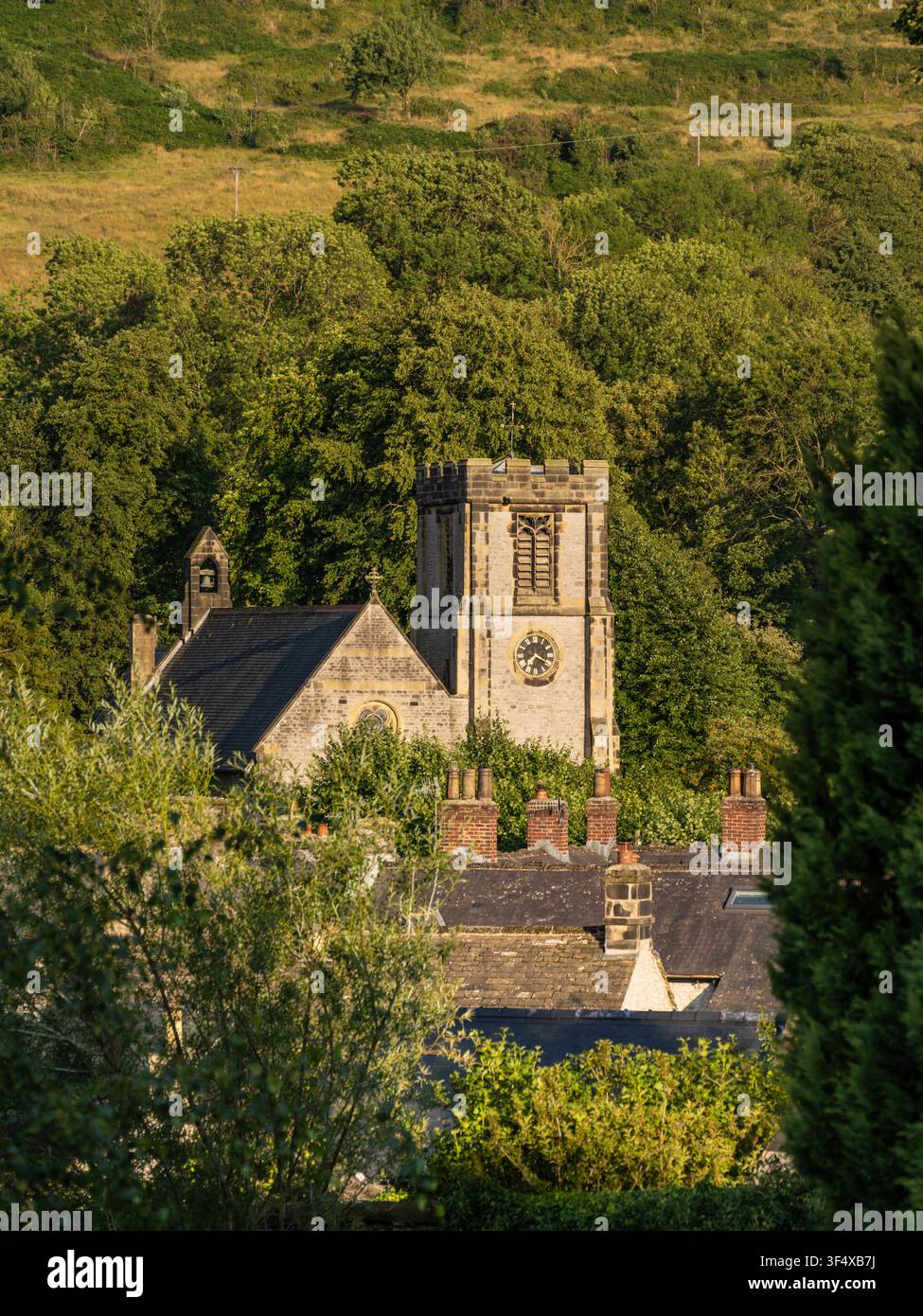 The historic stone tower of St Barnabas Church rises gracefully above village rooftops and lush green trees in Bradwell, Peak District, England Stock Photo