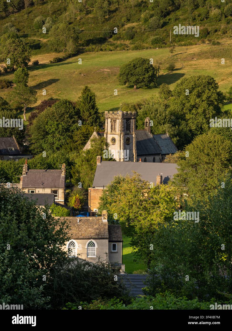 An elevated landscape view captures St Barnabas Church nestling among green trees and houses in the rural valley of Bradwell, Peak District, England Stock Photo