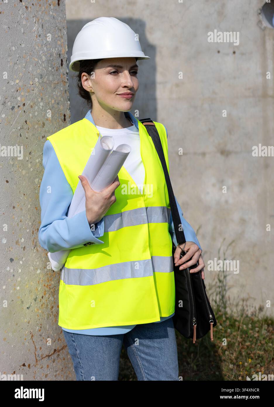 Woman engineer holding blueprints, wearing hard hat and safety vest at construction site Stock Photo