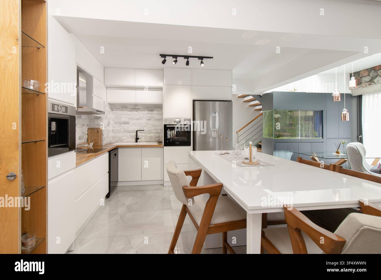 Modern open-plan kitchen and dining area featuring minimalist white cabinetry, stainless steel appliances, a large dining island with wooden chairs Stock Photo