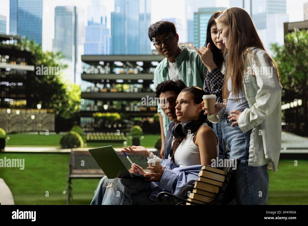 International students gather around laptop on campus bench, reviewing coursework notes and ideas together during outdoor learning collaboration. Group study project discussion. Stock Photo