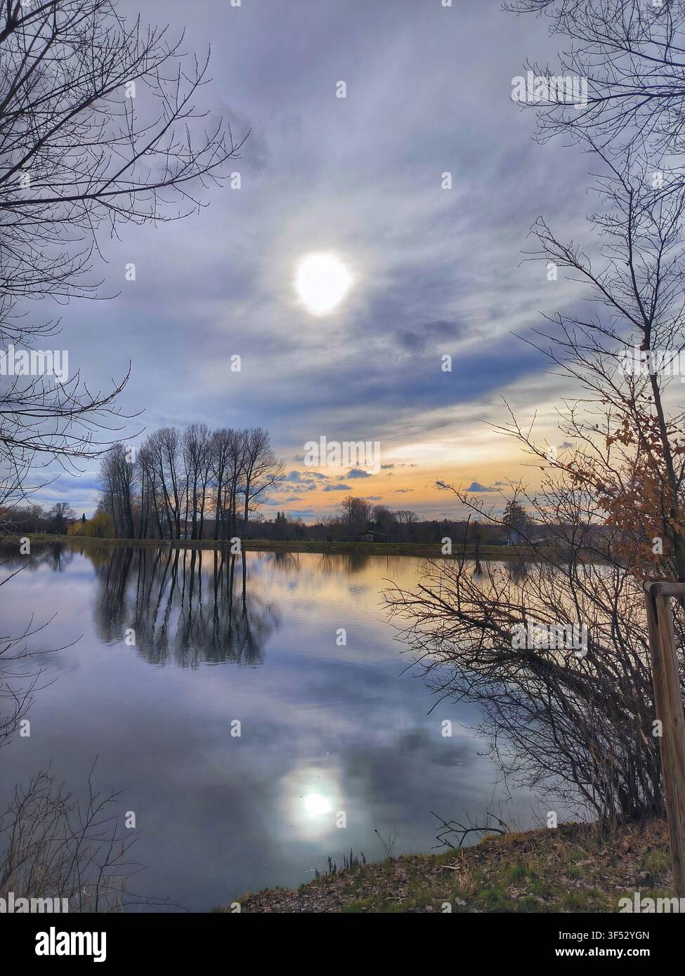 Beautiful scenic landscape of a calm lake reflecting cloudy sunset sky and silhouette of trees with the sun shining through clouds - Smartphone Captured Stock Image