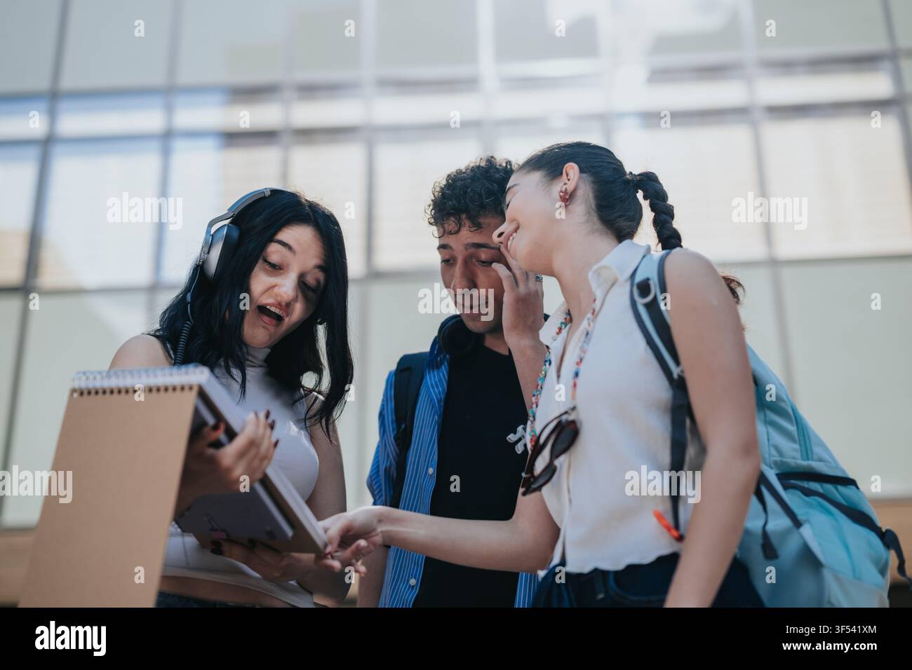 Three students studying together on campus sharing notes and a tablet while laughing and collaborating Stock Photo