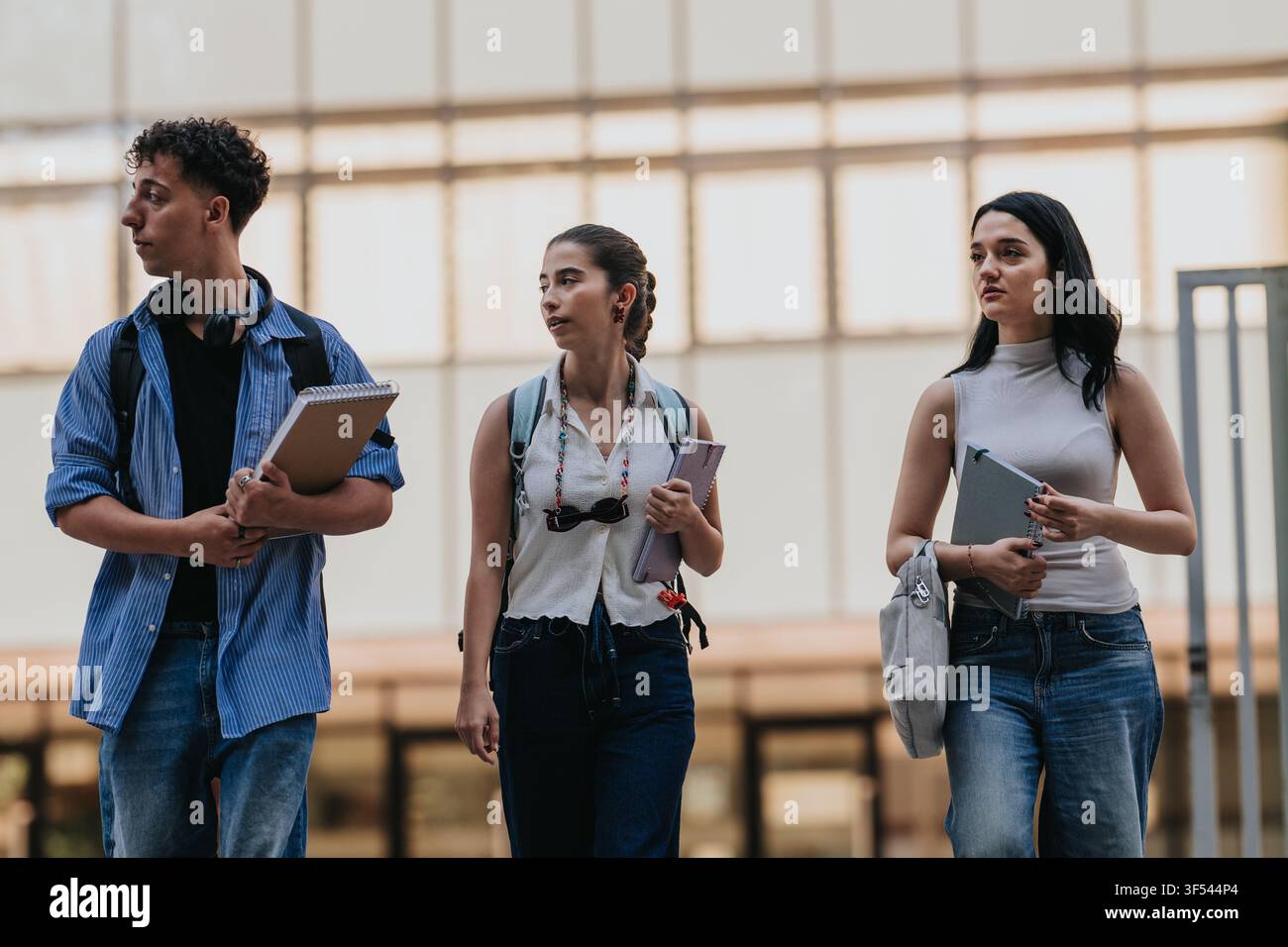 Three students walking on campus carrying notebooks and backpacks while talking between classes Stock Photo