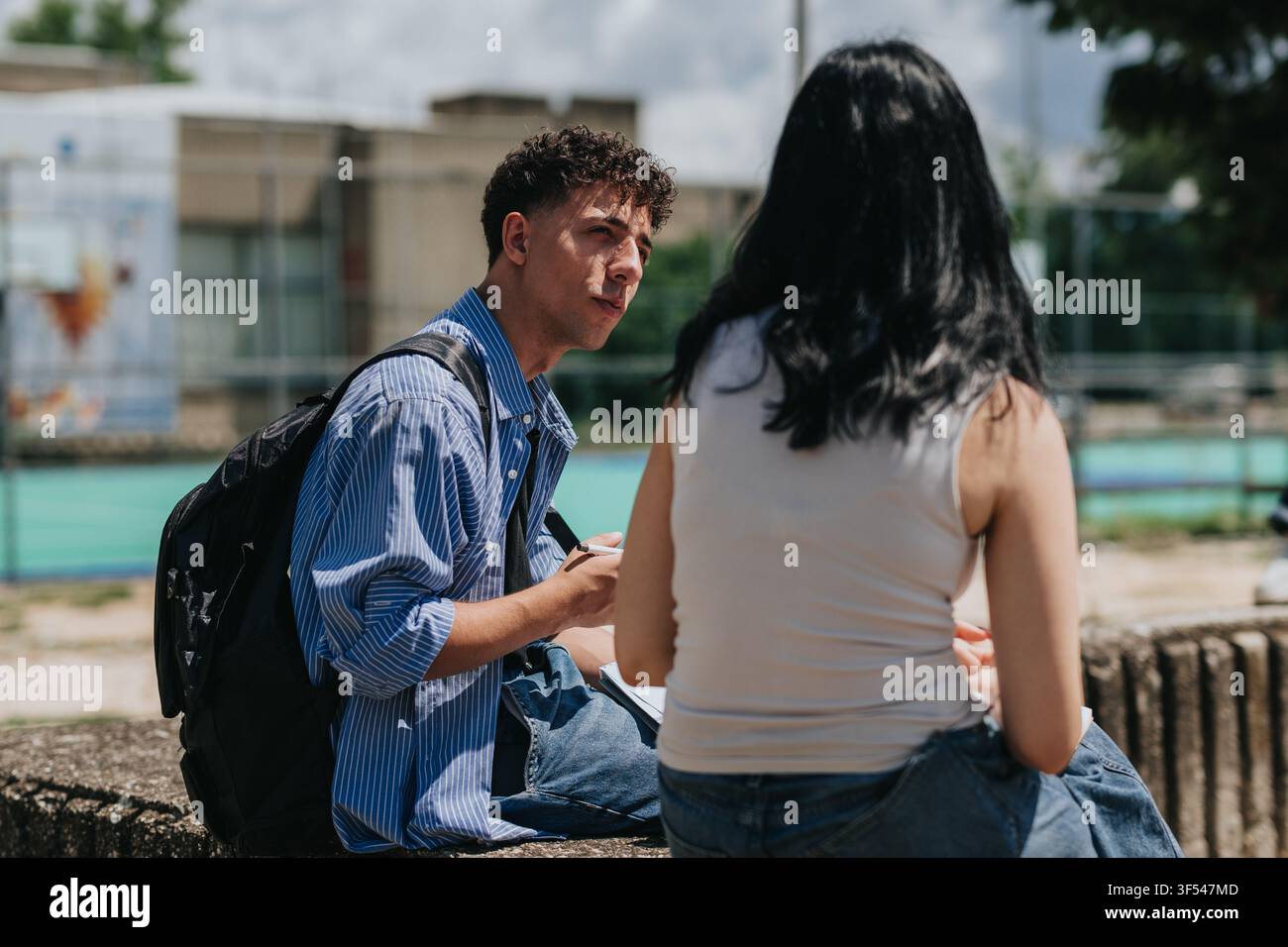 Two students talking outdoors on a campus bench with backpacks and notebooks Stock Photo