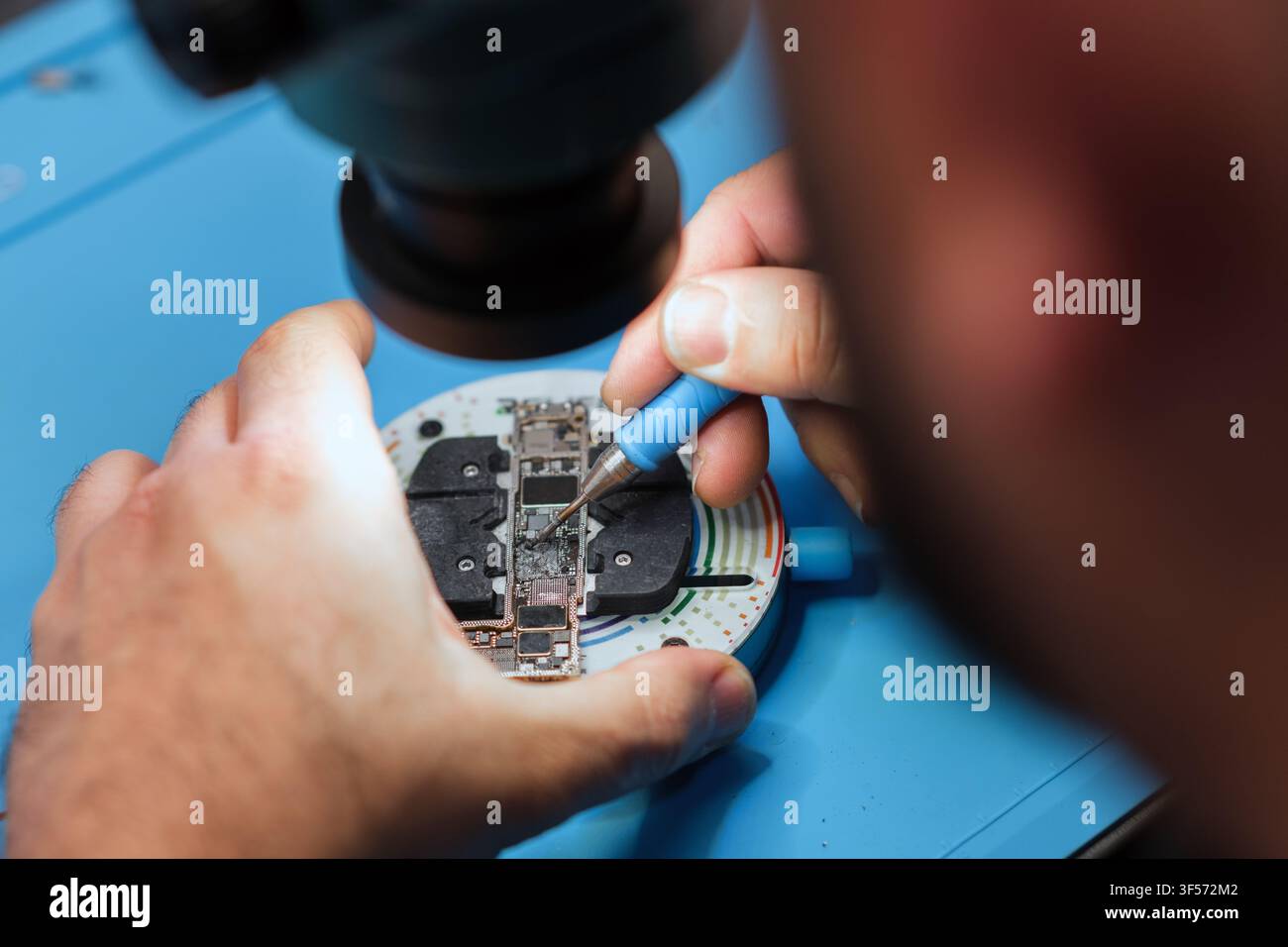 Technician repairs electronic circuit board with precision tools under magnifying lamp in a workshop setting Stock Photo