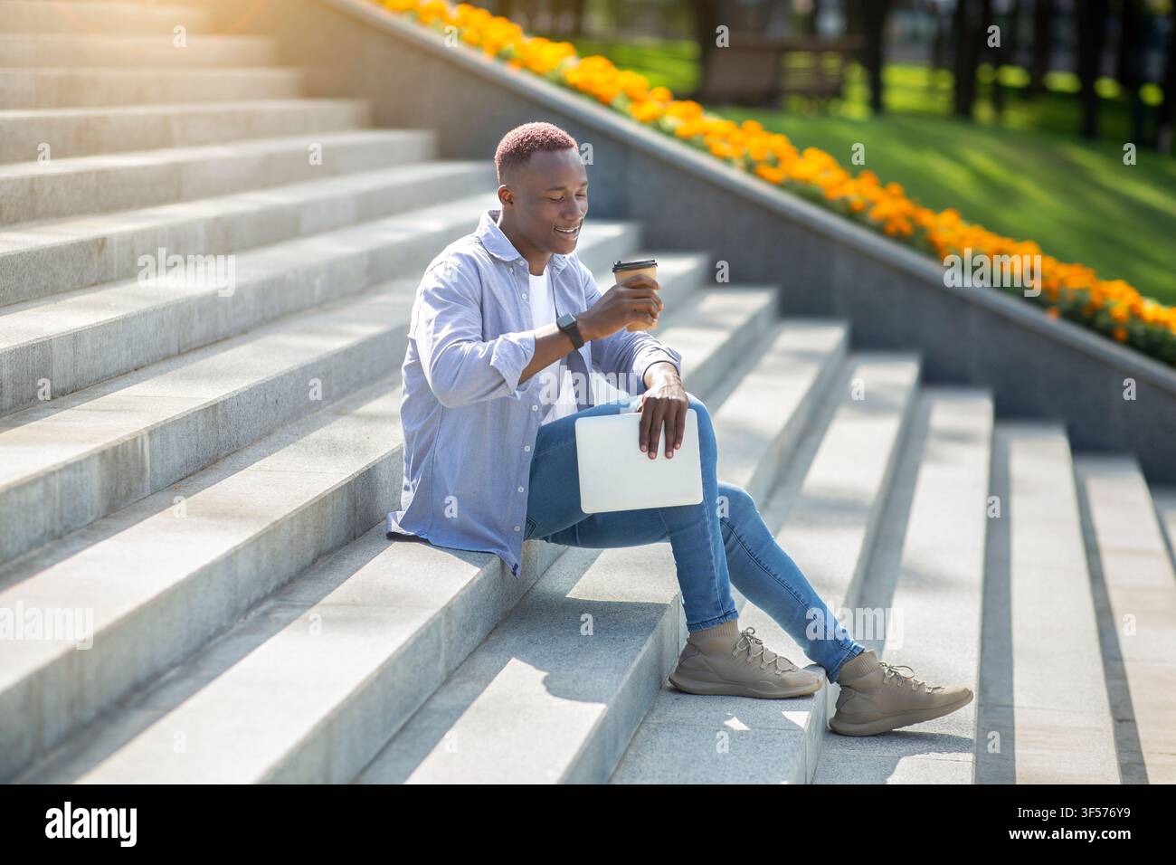Positive black guy with tablet computer drinking takeaway coffee on stone stairs downtown Stock Photo