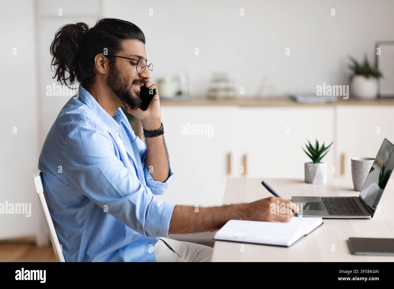 Cheerful Arab Freelancer Guy Talking On Cellphone And Writing Notes To Notepad Stock Photo