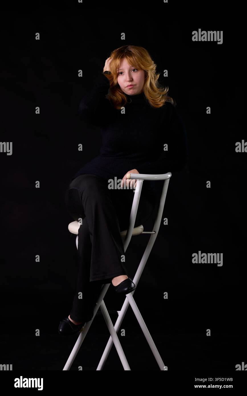 Young woman sitting on a white chair, looking pensive with a dark background Stock Photo