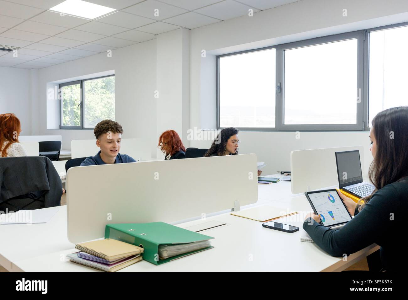 MBA students work independently and collaborate at desks in a bright modern university classroom, using laptops, tablets, notebooks, and study materia Stock Photo
