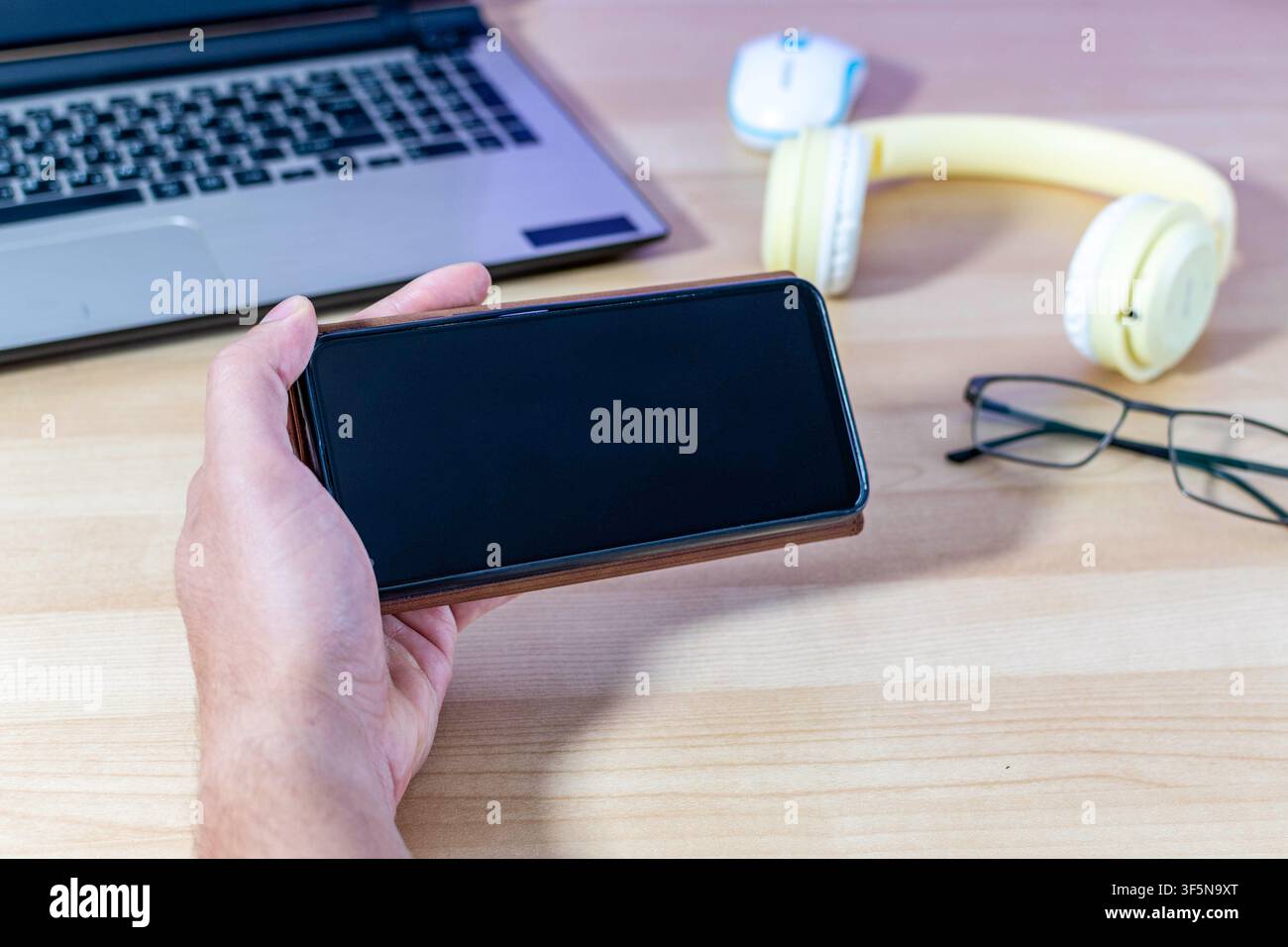 A man holds a mobile phone in a brown case in landscape mode over his desk. The black screen is ideal for mockups. Stock Photo